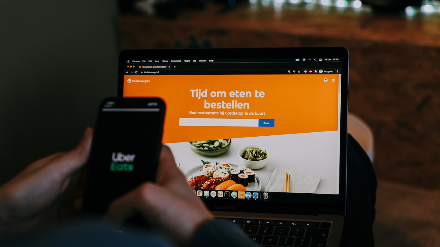 Hands hold smartphone with Uber Eats logo next to laptop.